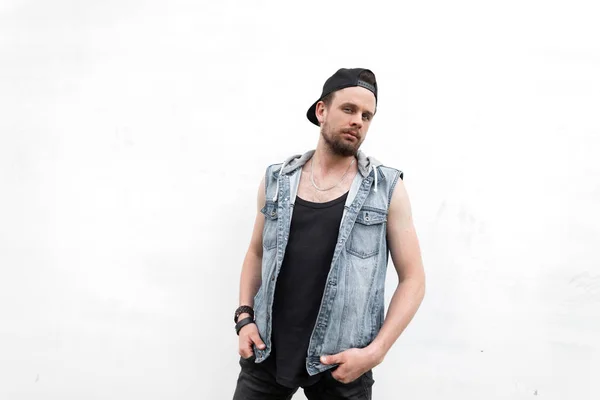 Jeune homme hipster américain avec une barbe dans un gilet en denim bleu à la mode dans un t-shirt dans un élégant chapeau noir se dresse près d'un mur vintage blanc dans la ville. Moderne mignon gars en plein air été . — Photo