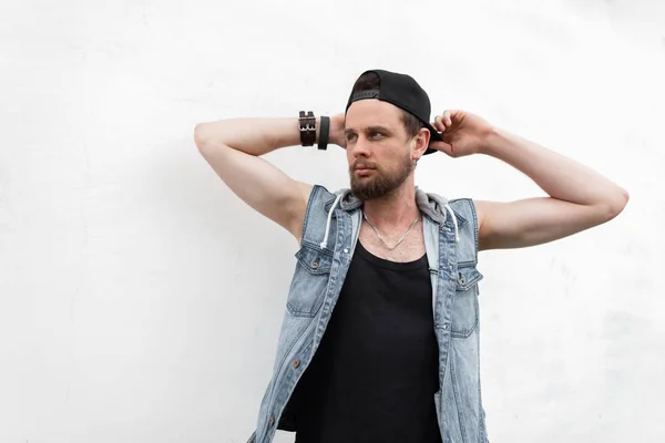 Brutal bonito jovem hipster homem com uma barba em um colete jeans azul vintage em uma camiseta preta posando em uma cidade perto de um edifício branco. American cara atraente ao ar livre dia de verão. Moda masculina . — Fotografia de Stock