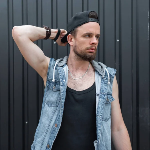 Nice young man hipster with a beard in a stylish black cap in a fashionable blue denim vest in a trendy T-shirt stands near a metal black wall. Handsome guy enjoys a warm summer day. Menswear.