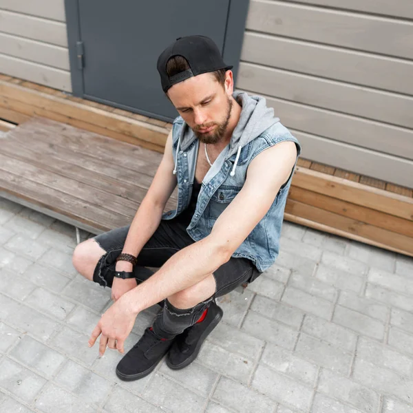 Jovem urbano moderno em roupas da moda em um elegante boné preto descansa sentado em uma escada de madeira perto de um edifício vintage na cidade. Um tipo bonito relaxa ao ar livre. Vista de cima . — Fotografia de Stock