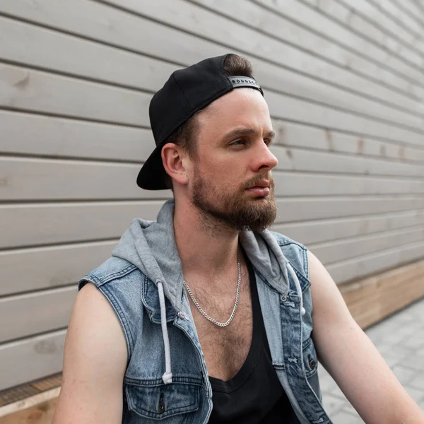 Jovem homem brutal hipster em um boné preto em um colete de ganga elegante em uma camiseta com barba goza o fim de semana sentado perto de um edifício de madeira na cidade. na moda urbano bonito cara na rua . — Fotografia de Stock