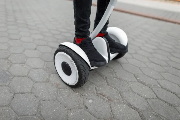 Fechar os pés de um homem num hoverboard moderno. Cara elegante em jeans em tênis de pé em um hoverboard branco na cidade em um dia de verão . — Fotografia de Stock