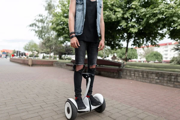 Jeune homme élégant en jean noir déchiré dans un gilet en denim dans un t-shirt en baskets à la mode se tient un hoverboard dans la rue par une journée ensoleillée d'été. Un citadin sur le tableau blanc à l'extérieur. Loisirs . — Photo