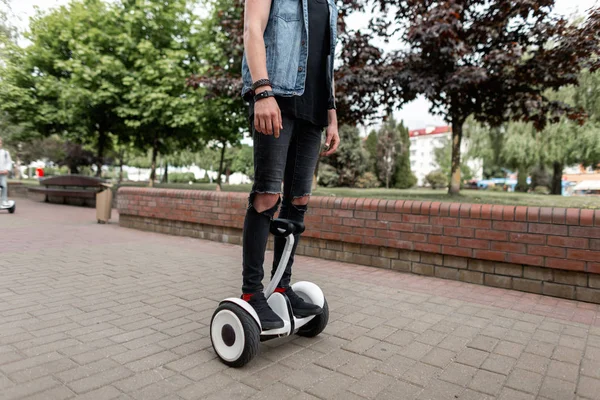 Giovane con un giubbotto di jeans in maglietta con jeans strappati e scarpe da ginnastica che cavalcano un girocollo bianco in fondo alla strada. I piedi degli uomini sull'hoverboard sullo sfondo della città. Piccolo fine settimana . — Foto Stock