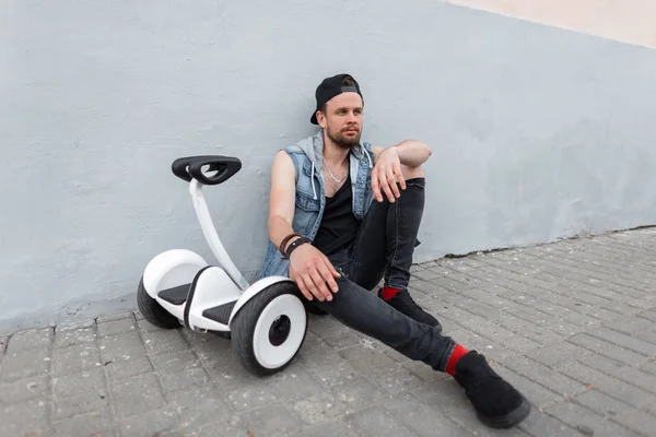 Élégant jeune homme dans un gilet en jean bleu vintage en jeans noirs en baskets dans une casquette à la mode se repose après une balade sur un hoverboard près d'un bâtiment de la ville. beau mec est relaxant . — Photo