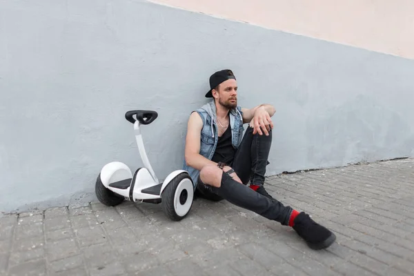 Madern gentil jeune homme dans un gilet en denim bleu vintage en jeans noirs en baskets dans une casquette à la mode se repose après une balade sur un hoverboard près d'un bâtiment dans la ville. élégant gars est relaxant . — Photo