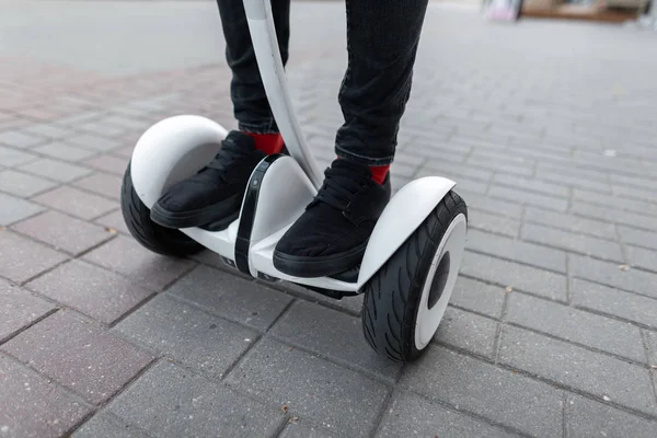 Pria bergaya jins dengan sepatu kets dengan kaus kaki merah mengendarai hoverboard putih di sekitar kota pada hari musim panas. Pendekatan kaki laki-laki dan giroskop listrik putih. Teknologi modern. Gaya hidup aktif . — Stok Foto