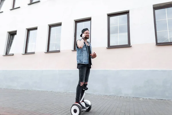 Giovane bell'uomo in jeans neri strappati in un gilet di jeans con un berretto in una t-shirt in scarpe da ginnastica su un hoverboard in giro per la città e parlando al telefono in una giornata estiva soleggiata. Ragazzo urbano sulla lavagna bianca . — Foto Stock