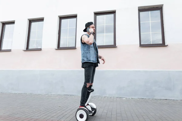 Attraente giovane uomo in un giubbotto jeans strappato in un berretto in una t-shirt in scarpe da ginnastica cavalca su un giroscopio bianco e parla su un telefono cellulare.Ragazzo elegante viaggia per la città in piedi su hoveybord . — Foto Stock