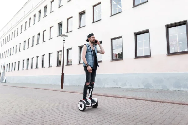 American young man in ripped stylish jeans in a denim vest in a cap in a t-shirt in sneakers on a hoverboard around the city and talking on the mobile phone. Nice guy on the white gyroscope outdoors. — Stock Photo, Image