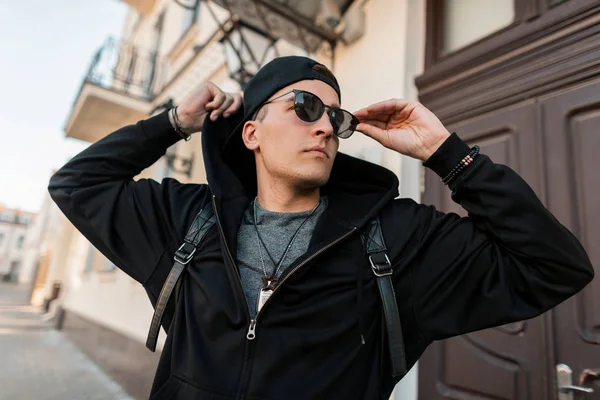 Americký mladý hipíků ve stylových černých šatech v módní čepici v brýlích s stylovým batohu po celém městě za slunného letního dne. Atraktivní městský model má rád víkend. — Stock fotografie
