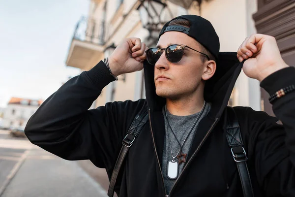 Elegante hombre hipster joven con una sudadera con capucha en una gorra de béisbol en gafas de sol con una mochila está posando cerca de un edificio vintage en un día soleado. Moderno buen chico viajar por las calles de la ciudad . — Foto de Stock