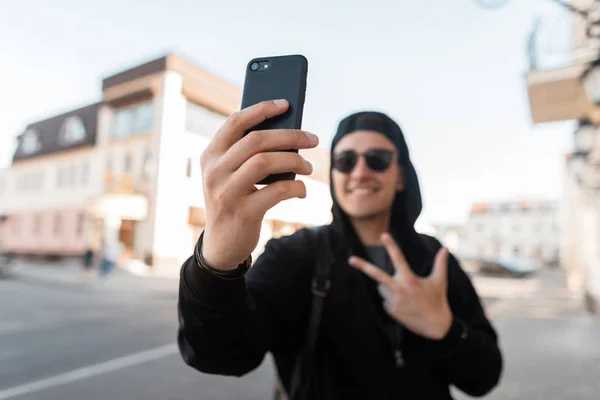 Cheerful young hipster man in hoodie in sunglasses in trendy baseball cap is smiling and showing peace sign making selfie on phone. Positive handsome guy walks down the street near vintage buildings.