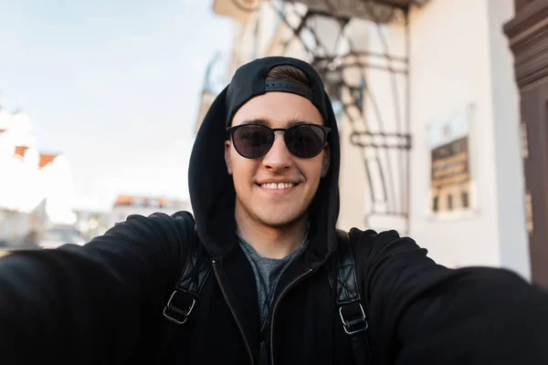 Homem hipster alegre jovem em camisola de capuz preto na moda com um gorro em óculos de sol sorri e tira fotos de si mesmo no telefone. Modelo cara positivo anda pela cidade em um dia ensolarado . — Fotografia de Stock