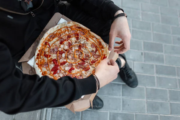 Young man in black clothes is holding a box of hot delicious pizza with sausage with tomatoes and cheese dressing with spicy seasonings. Guy took a break for lunch. Fast tasty food. Close-up.