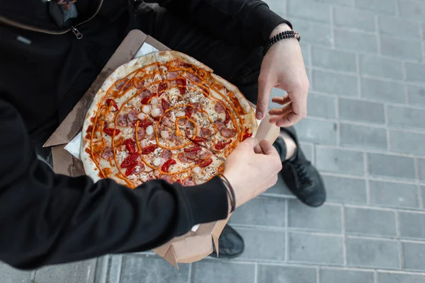 Siyah elbiseli genç adam elinde sosisli, domatesli ve baharatlı peynirli bir kutu sıcak lezzetli pizza tutuyor. Adam öğle yemeği molası verdi. Lezzetli yemekler. Yakın plan..