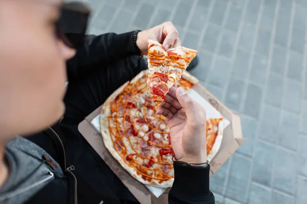 Stylish young man in sunglasses holding a slice of delicious hot pizza. Guy is sitting on the street and eating pizza. Dinner time. Close-up.