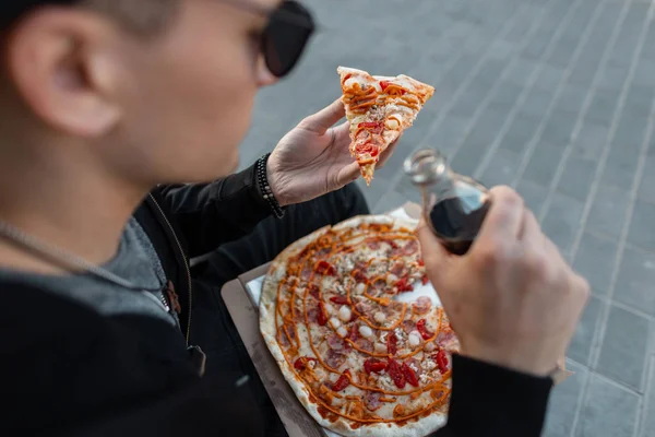 Joven hipster de moda en gafas de sol oscuras con una rebanada de sabrosa pizza caliente en la calle. El guapo está sentado en la ciudad comiendo pizza. Hora del almuerzo. Primer plano . — Foto de Stock