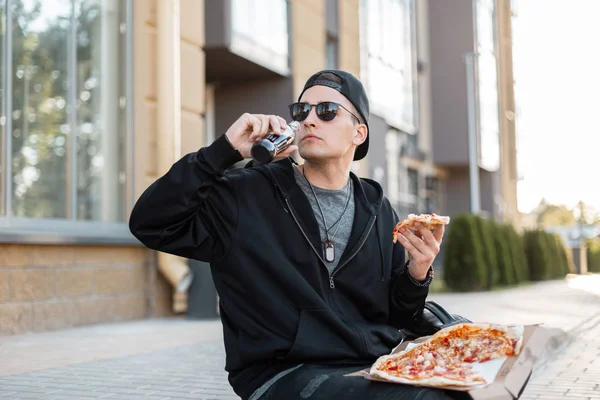 American young hipster man in fashionable black clothes in a trendy cap in stylish sunglasses with a hot pizza sits on the street in the city. Handsome modern guy drinks soda at lunchtime. Dinner.