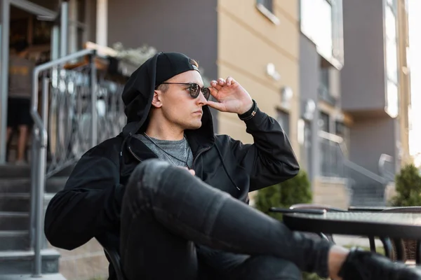 O jovem hipster americano na roupa preta elegante em óculos escuros na moda em um gorro de beisebol na calça senta-se em um café de verão na rua em um dia ensolarado de verão. Bonito cara está descansando ao ar livre . — Fotografia de Stock