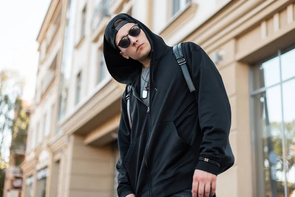 Homem hipster americano jovem em um gorro na moda em uma camisola preta em óculos de sol elegantes com uma mochila posa na rua perto de um edifício vintage. Um tipo porreiro viaja. Moda de rua moderna . — Fotografia de Stock