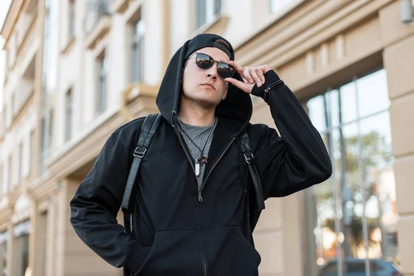 Jonge Amerikaanse hipster in een stijlvolle sweatshirt met capuchon en donkere zonnebril in een pet met rugzak staat in een stad vlakbij een modern gebouw. Leuke koele kerel loopt door de straat in de zomer dag. — Stockfoto