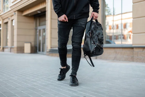 Bir aslan desenli bir deri sırt çantası ile vintage kot şık bir siyah sweatshirt şık genç adam binaların yakınındaki cadde boyunca yürüyor. Modern adam dışarıda. Close. Erkek giyim