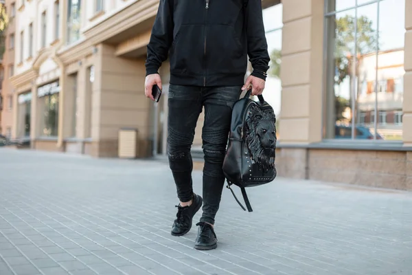 Stijlvolle jonge man in een trendy zwarte sweater met een lederen zwarte rugzak met een leeuw patroon in jeans loopt door de straat in de stad. Nieuwe collectie modieuze herenkleding. Close-up. — Stockfoto