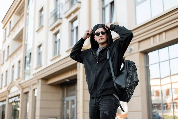 Αστικός νεαρός χίπης σε ένα μοντέρνο φούτερ με κουκούλα με γυαλιά ηλίου σε ένα μαύρο καπάκι με ένα κομψό σακίδιο σε τζιν ταξιδεύει γύρω από την πόλη σε μια φωτεινή καλοκαιρινή μέρα. Σύγχρονος τύπος στο δρόμο. — Φωτογραφία Αρχείου