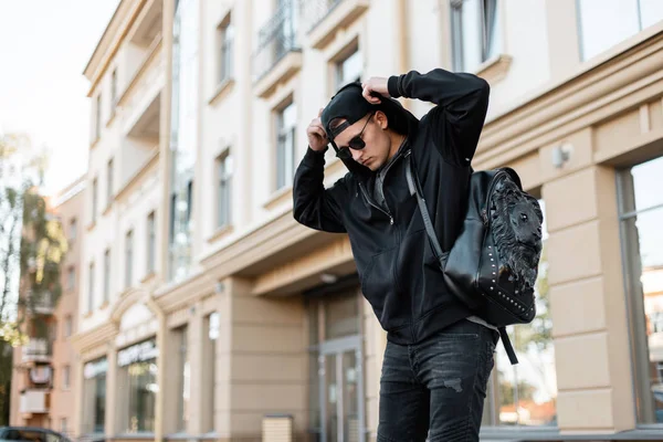 Młody amerykański hipster w modnej czapce w czarnej bluzie w stylowych okularach przeciwsłonecznych w dżinsach ze skórzanym plecakiem prostuje kaptur w pobliżu nowoczesnego budynku. Miejski facet podróżuje po mieście.. — Zdjęcie stockowe