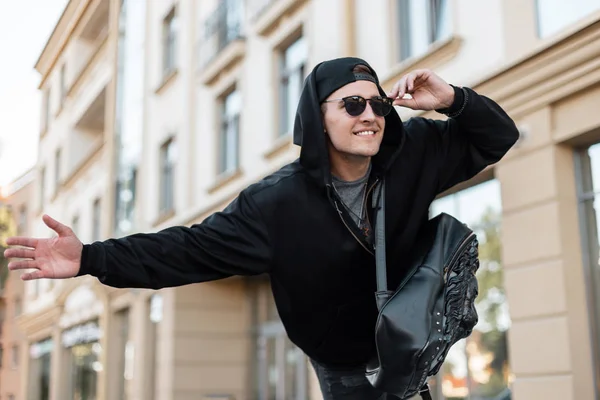 Mladý šťastný pohledný muž hipster ve stylovém oblečení v černé čepici s módním koženým batohem v tmavých slunečních brýlích ve městě. Radostný chlapík chytající taxi na ulici. — Stock fotografie