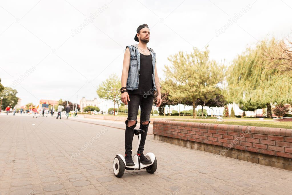 Young handsome man in ripped black jeans in a denim vest in a cap in a t-shirt in sneakers rides a hoverboard through the city on a summer sunny day. Urban guy on the white board. Leisure. 