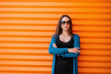 Vintage kot bir örme yaz pelerin siyah bir t-shirt güneş gözlüğü Pretty güzel genç hipster kadın şehirde metalik turuncu duvar yakınında duruyor. Şık şık kız modeli.