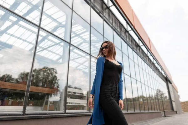 Elegante jovem hipster em uma capa de malha azul vintage em jeans elegantes em uma camiseta com renda em óculos de sol da moda caminha pela cidade em um dia de verão. Menina bonita perto do edifício de vidro . — Fotografia de Stock