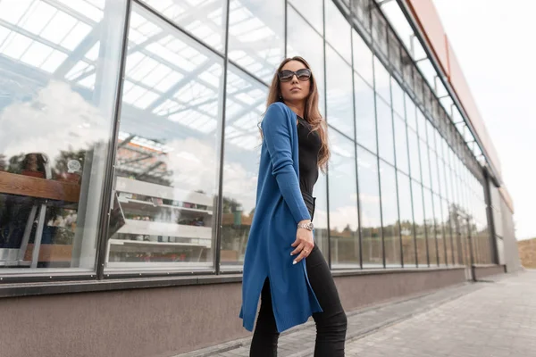 Mujer hipster joven con estilo en una capa larga de punto azul en una parte superior en jeans con gafas de sol camina por la ciudad cerca de un edificio de vidrio. Chica urbana moderna viaja por la calle en un día de verano . —  Fotos de Stock