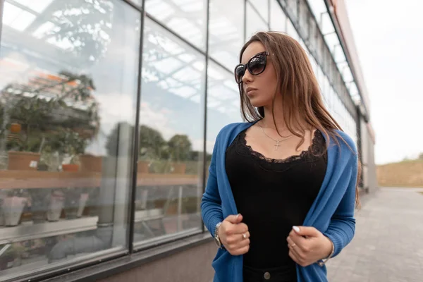 Urban joven mujer bastante hipster en una capa azul de moda de punto en gafas de sol de moda en una camiseta de color negro vintage está de pie cerca de un edificio de vidrio. Elegante modelo de chica descansando. Estilo de verano juvenil . —  Fotos de Stock