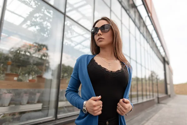 Całkiem efektowna młoda kobieta Hipster w stylowe okulary przeciwsłoneczne w czarnym T-shirt z koronką w niebieskim dzianiny Cape stoi w pobliżu szklany budynek na zewnątrz. Atrakcyjna dziewczyna spacerów po mieście. Letnich. — Zdjęcie stockowe