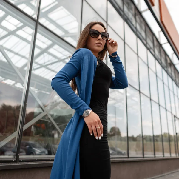 Joven hipster glamurosa en una capa de punto largo vintage en jeans en una camiseta con gafas de sol de moda camina por la calle cerca del edificio de cristal. Atractiva chica bastante elegante en la calle . —  Fotos de Stock