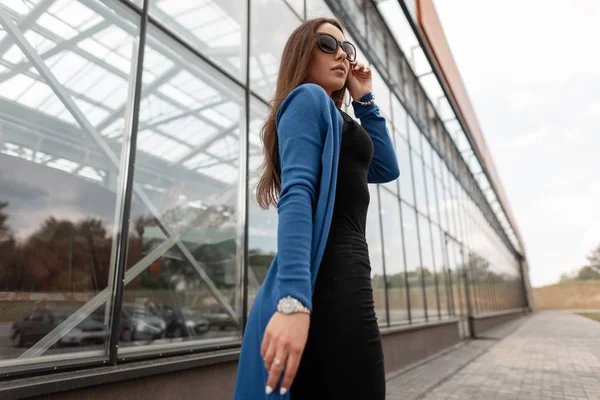 Mujer hipster joven bonita urbana en gafas de sol en una capa de punto vintage en una camiseta en jeans con estilo camina en un día de verano cerca de un edificio de vidrio moderno en la ciudad. Hermosa chica de moda . —  Fotos de Stock