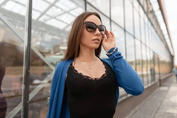 Mulher bonita nova do hipster na capa de malha azul nos óculos de sol na moda na t-shirt elegante preta está de pé perto do edifício de vidro na rua. Luxuosa menina bonita relaxa. Moda de verão . — Fotografia de Stock