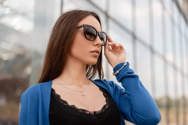 Retrato de una hermosa mujer hipster joven con labios sexy en una capa elegante azul de punto en una camiseta elegante negro con encaje cerca del edificio de vidrio moderno en la ciudad. Chica atractiva al aire libre . — Foto de Stock