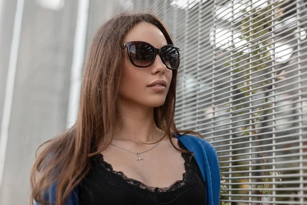 Jeune et glamour jolie femme hipster en cape bleue tricotée en lunettes de soleil à la mode dans un t-shirt noir se tient près d'un mur métallique d'une rue de la ville. Modèle fille mignonne en plein air. Style d'été . — Photo