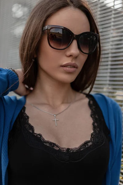 Ziemlich attraktive junge Hipster-Frau mit trendiger dunkler Sonnenbrille in einem schwarzen Vintage-T-Shirt mit Spitze in einem Strickcape posiert in der Nähe einer Metallwand in der Stadt. Porträt eines schönen Mädchens. Sommer-Stil. — Stockfoto