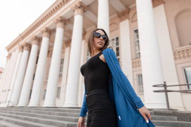 Şık örme uzun pelerinli çekici genç hipster kadın güneş gözlüğü ile kot pantolon bir üst sütunlar ile vintage bir binanın yakınında şehir etrafında yürüyor. Sokakta seyahat güzel kız.