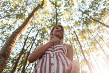 Şık pembe çizgili elbise pretty mutlu genç kadın ormanda duruyor ve parlak güneş ışığı sahiptir. Sevimli bir gülümseme ile neşeli kız parkta yürüyor. Modaya uygun kadın yazlık elbiseleri. Alt görünüm