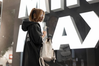Modaya uygun deri beyaz sırt çantası ile vintage gözlük bir trendy uzun ceket bir saç modeli ile Modern genç kadın cam duvarın yakınında mağaza etrafında yürüyor. Tatilde Kentsel çekici kız.