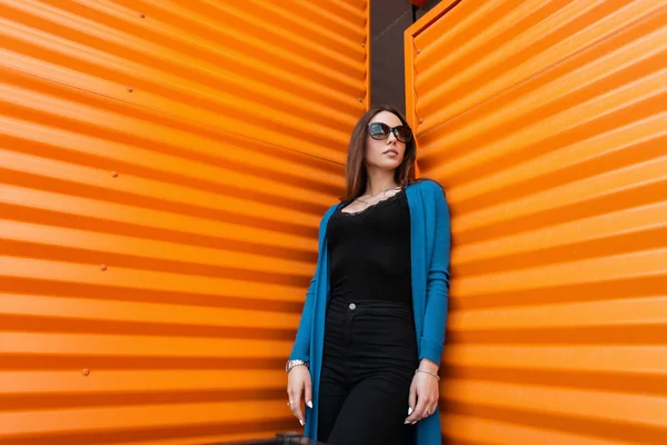 Fashionable pretty young hipster woman in a trendy knitted cape in jeans in a t-shirt in stylish sunglasses stands near a bright orange metal wall in the city. Attractive girl outdoors summer day.