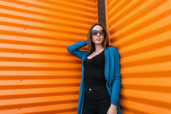 Stylish beautiful young hipster woman in sunglasses in a black T-shirt in a knitted summer cape in vintage jeans is standing near the metallic orange wall in the city. Fashionable pretty girl model.