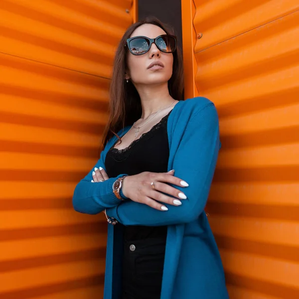 Ganska söt hipster ung Urban kvinna i trendiga kläder i snygga solglasögon poserar utomhus nära en ljus orange metall vägg. Sexig glamorösa Girl Fashion Model. Trendiga damkläder. Stil. — Stockfoto