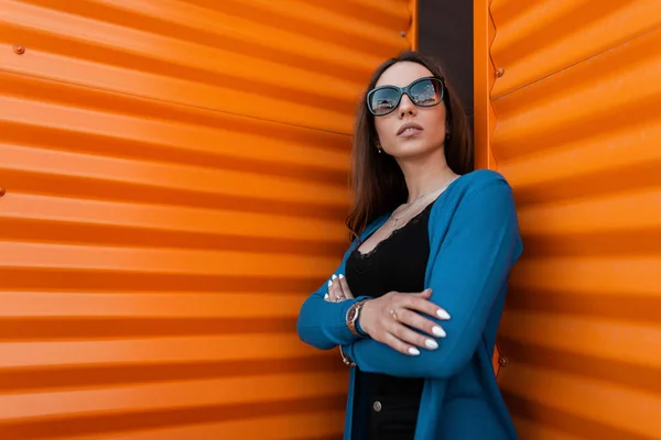 Atractiva mujer joven hipster en una capa de punto largo de moda en un top negro con estilo en jeans negros en gafas de sol vintage posa cerca de una pared de metal en la calle. Hermosa chica relaja al aire libre . —  Fotos de Stock